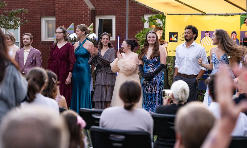 A group of singers in formal wear stand on an outdoor stage in front of a crowd.