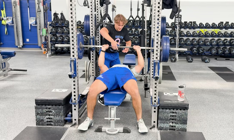 Denison student assists weightlifter with a rep on a weight bench in a gym.
