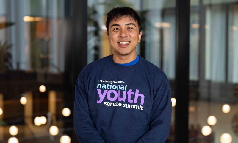Photo of Yahir Fernandez-Alvarez ’26 wearing a National Youth Service Summit shirt, standing in front of a large glass wall inside a modern building.