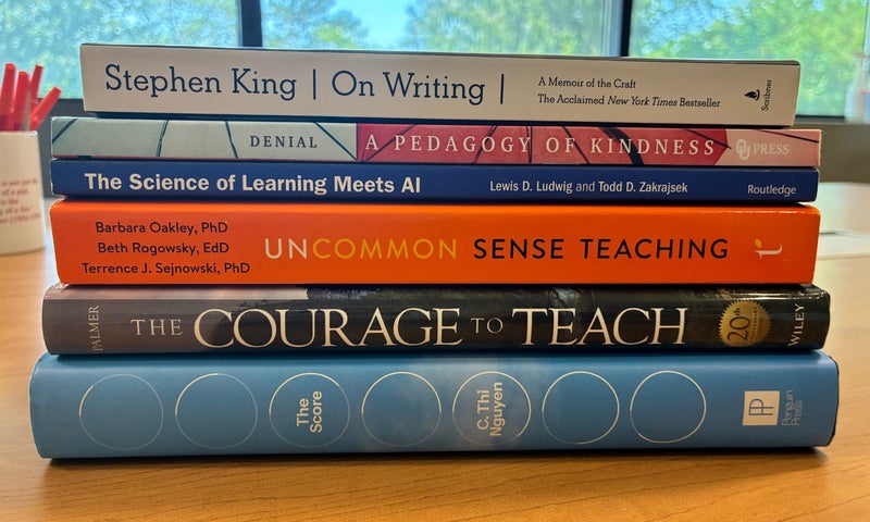 Five books stacked on a table, including On Writing by Stephen King and titles about AI, pedagogy, teaching, and learning, with a window view of trees in the background.