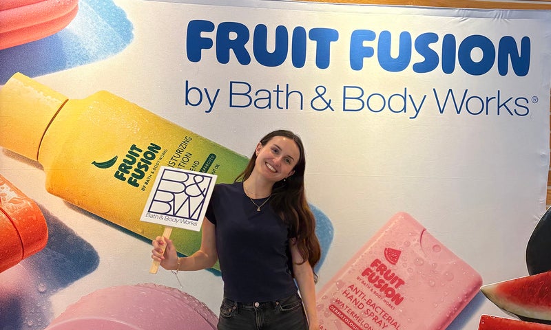 A person stands in front of a Bath & Body Works Fruit Fusion display, holding a sign and smiling at the camera.
