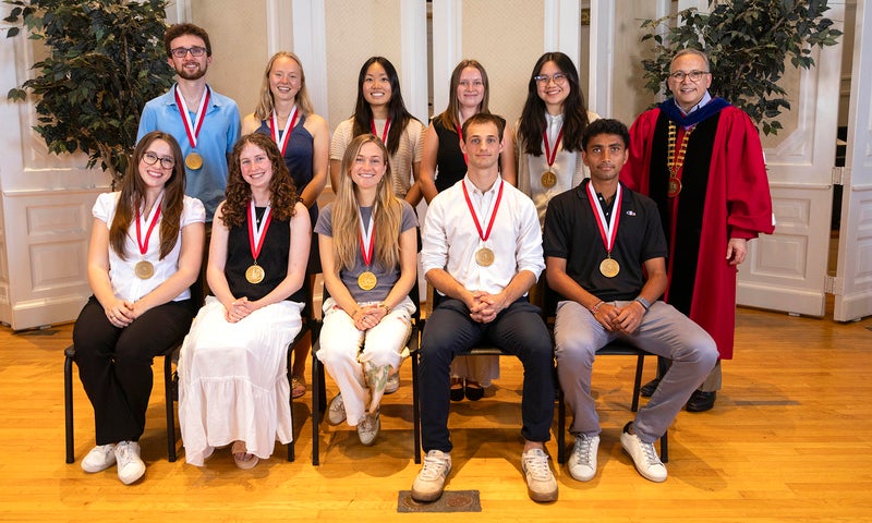 A group of ten Denison students, all 2026 President's medalists with medals, and President Adam Weinberg in academic regalia pose indoors, with plants and wood flooring in the background.