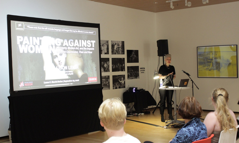 A speaker stands at a podium in front of a projection screen that reads Painting Against Women.