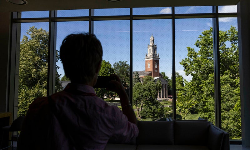 A person takes a photo of a college campus clock tower through large indoor windows on a sunny day.