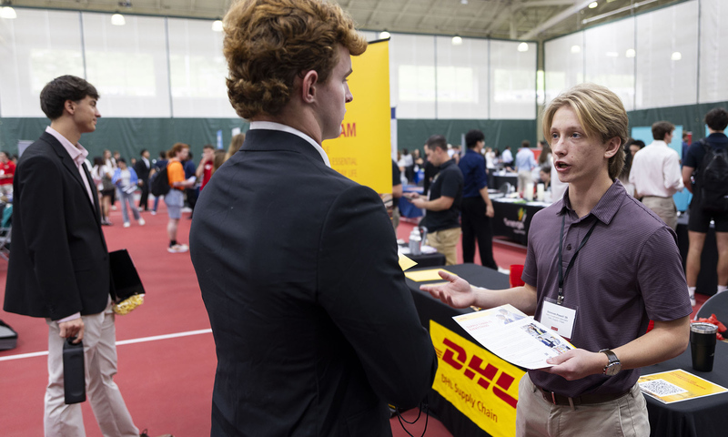 Two people speak at a career fair booth with a visible DHL sign, while others interact in the background inside a large gymnasium.
