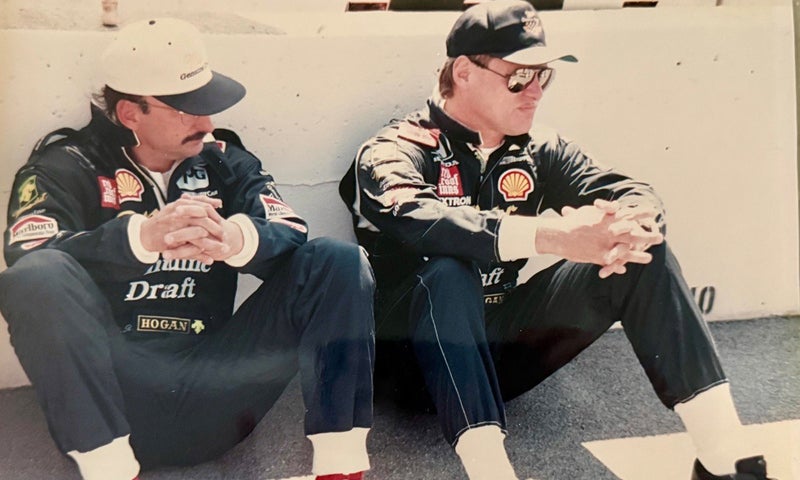 Two men in racing uniforms and caps sit on the ground against a wall, looking forward with hands clasped over their knees.