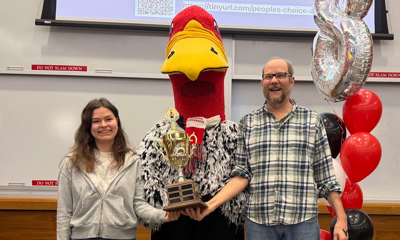 A young woman and man stand with a tall person in a buzzard mascot costume holding a trophy.