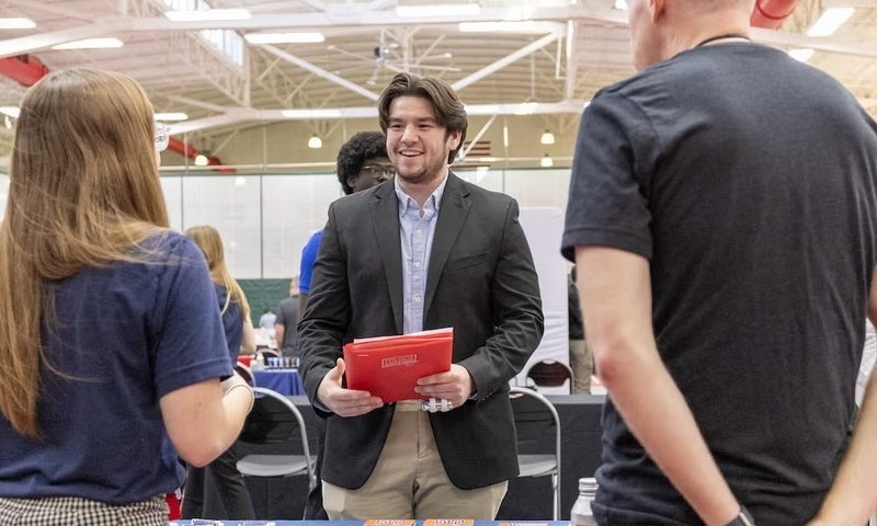 A man in a blazer holding a red folder talks to two people at a table during the career expo.
