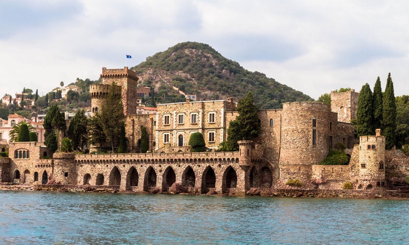A historic stone castle with arched walkways and towers sits beside a lake, with a wooded hill rising in the background under a cloudy sky.