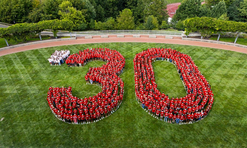 A large group of people wearing red shirts stand on grass arranged to form the number 30, with a few people in white shirts forming a dot before the 3. Trees and a path are in the background.