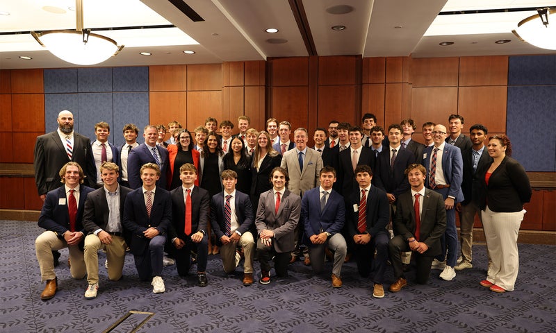 A large group of people in business attire pose for a group photo in a conference room with wood paneling and blue carpeting.
