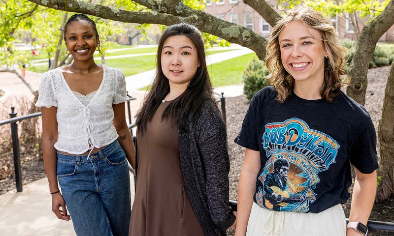 Three young women stand outdoors on a sunny day, posing and smiling in front of a leafy tree and campus buildings.