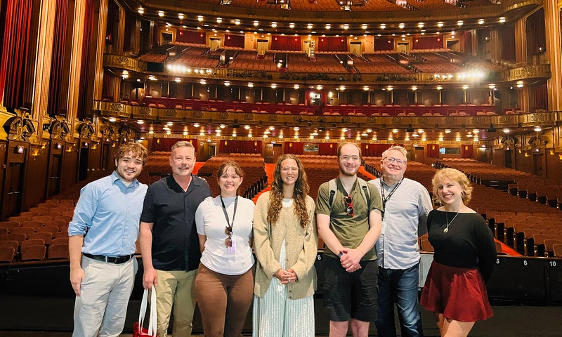A group of seven people stand on a theater stage with rows of empty red seats and ornate balconies visible behind them.