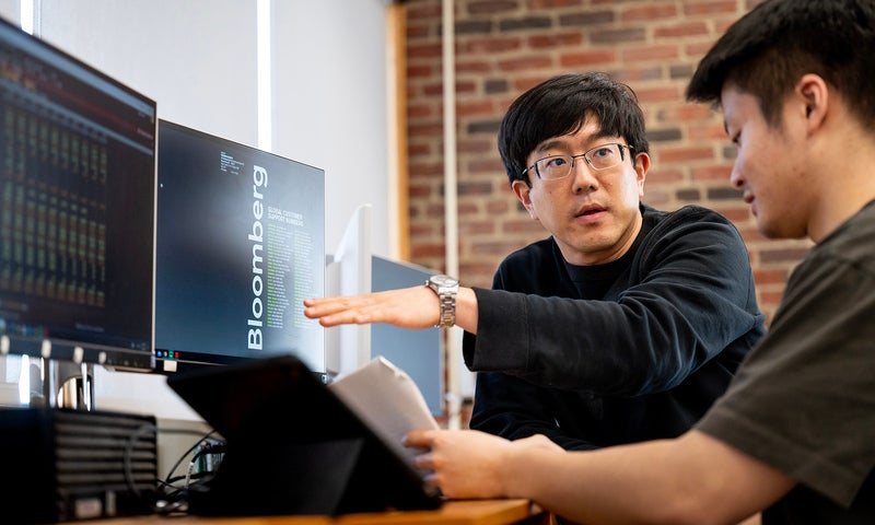 Two people work at a desk with computer monitors and a tablet, discussing data displayed on the screens in a modern office with exposed brick walls.