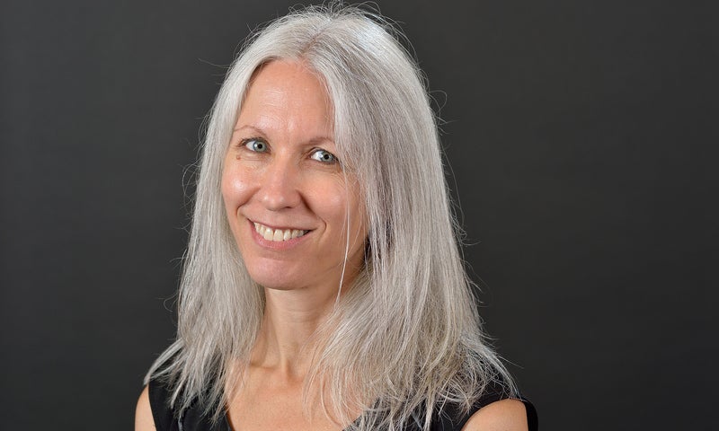 A woman with long, straight gray hair and a black sleeveless top smiles in front of a plain dark background.