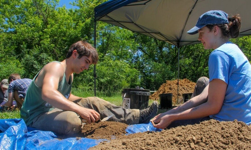Two people sit on the ground under a canopy, sifting through dirt on a blue tarp, working on an outdoor excavation or archaeology project (Photo credit: Allie Vugrincic)