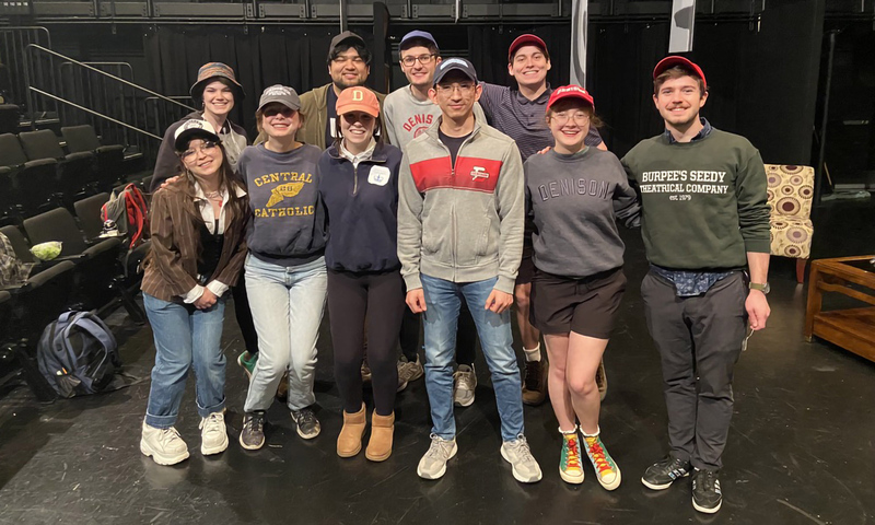 A group of ten young adults stand together on a stage, most wearing baseball caps.