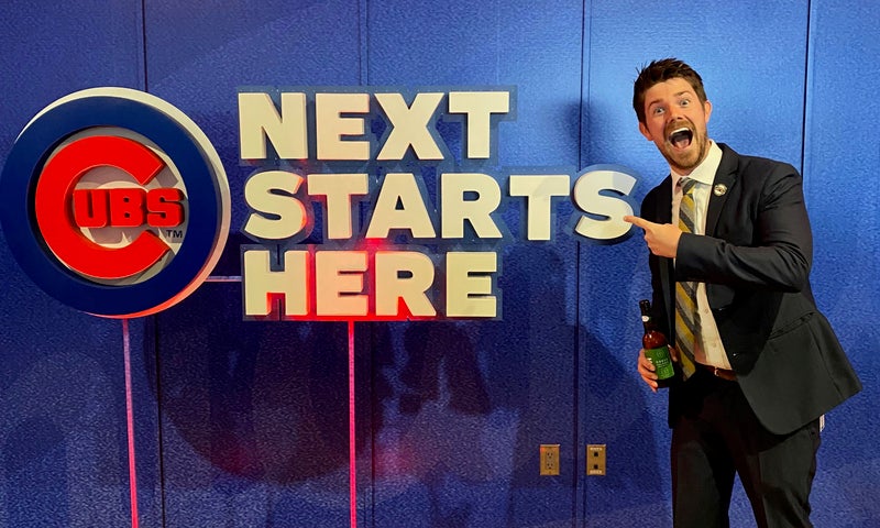 A man in a suit holding a drink points and smiles at a large sign that reads "Cubs NEXT STARTS HERE" on a blue wall.