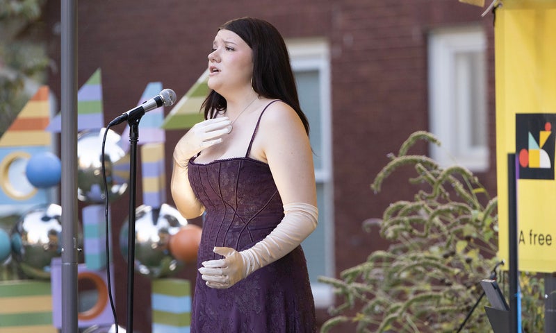 A woman with dark hair, wearing a purple sleeveless dress with straps, sings on an outdoor stage.