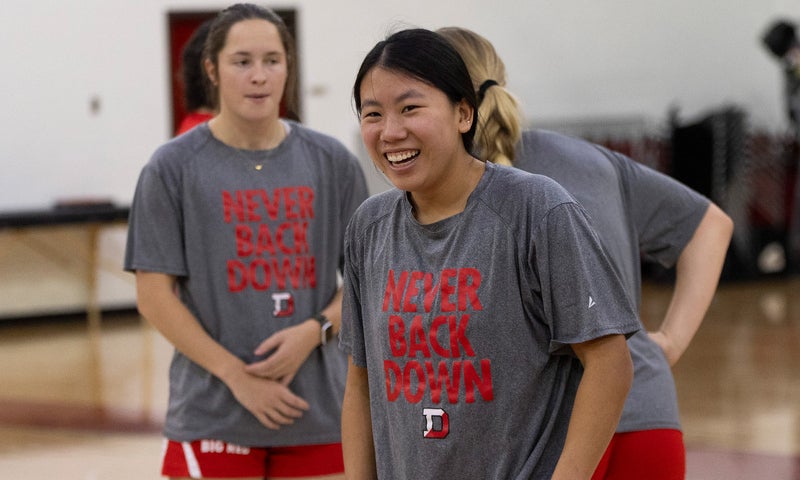 A women's basketball players in a Denison tee shirt that says "Never back down" smiles at practice with other players in the background.