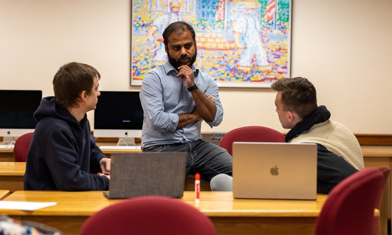 A man sits on a desk speaking with two students in a classroom with computers and colorful artwork on the wall in the background.