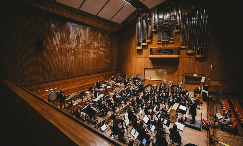 A wind ensemble seated in performance in a large concert hall flanked by projected images on a wood wall on one side and a large pipe organ on the other.