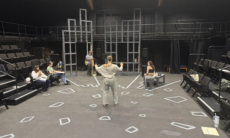 A person stands in the center of a theater stage marked with tape, facing four others seated or standing around the set, which includes geometric wooden frames and scattered chairs.