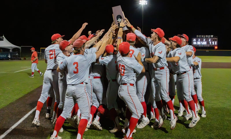 A team of baseball players grouped together on a baseball field at night hold up a championship trophy in celebration.