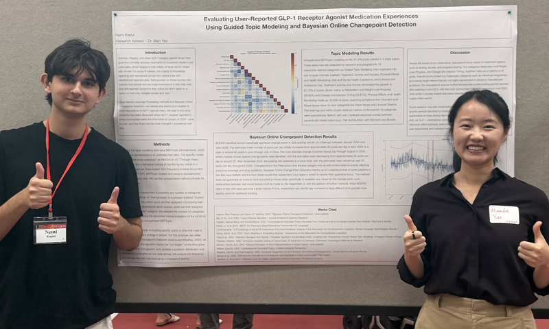 Two people stand in front of a scientific poster, both smiling and giving thumbs up, wearing name tags.