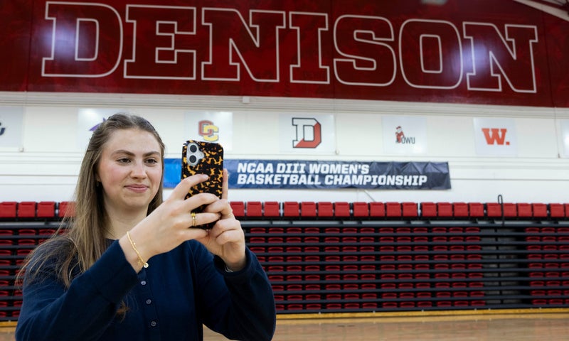 A person uses a phone to record a video inside a gymnasium with DENISON in large letters on the wall and a banner for the NCAA DIII Women's Basketball Championship in the background.