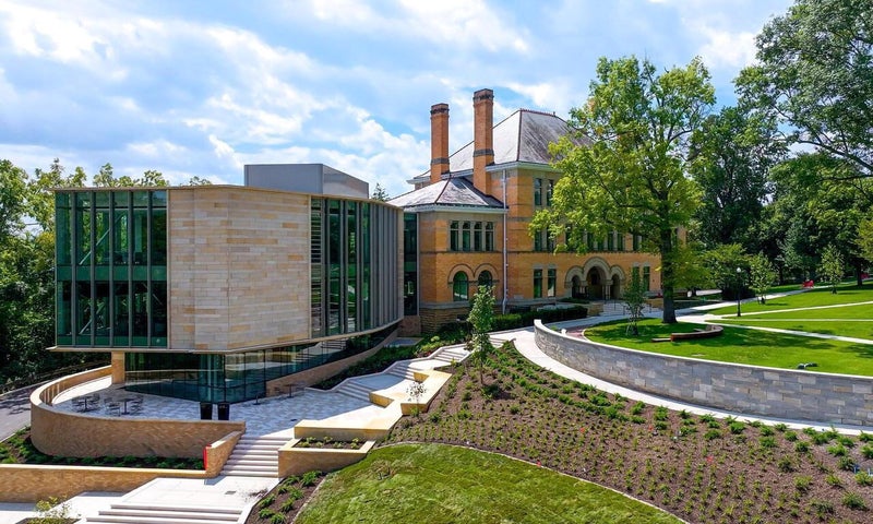 A modern building extension with glass and stone sits beside a large historic brick house, surrounded by green lawns, trees, and landscaped pathways under a partly cloudy sky.