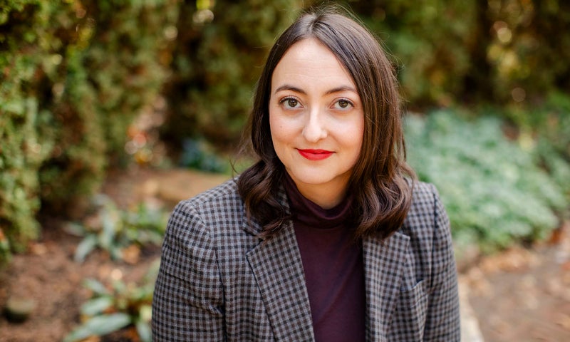 Dr. Krista Dalton with shoulder-length dark hair, wearing a checked blazer and dark top, sits outdoors in front of greenery, looking at the camera with a slight smile.