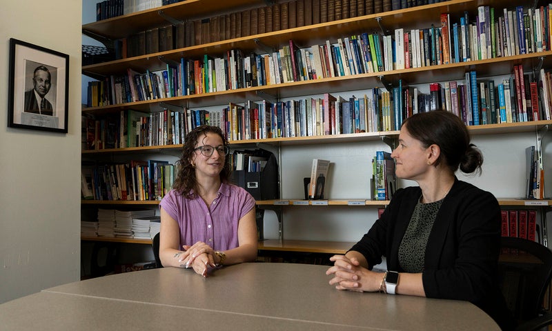 Two women having a conversation at a table in front of a wall of books; a black and white portrait of Denison alum Kirtley Mather hangs on the wall nearby.