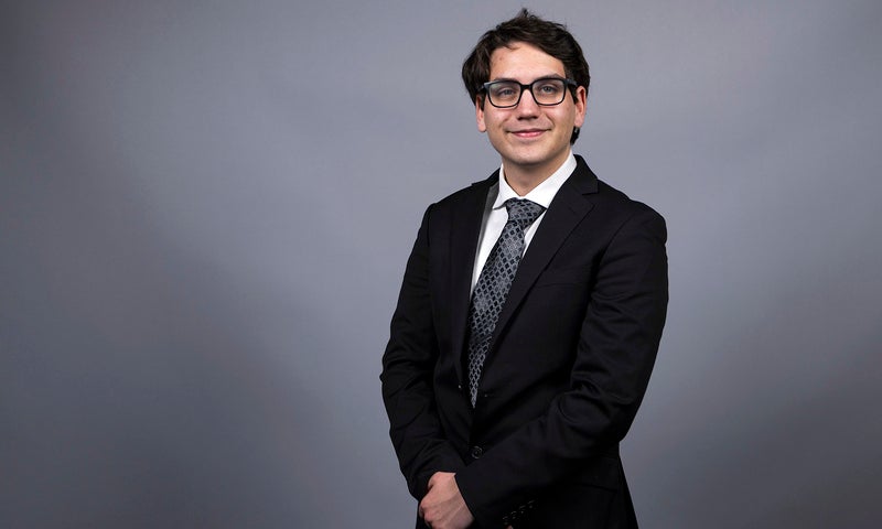 Max Levine ’26 wearing a dark suit and tie standing in front of a studio portrait backdrop.