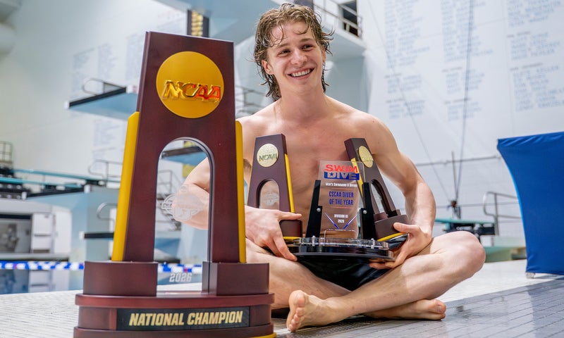 A young man in a swimsuit sits on a pool deck, smiling and holding several NCAA diving trophies, with a large “National Champion” trophy in the foreground and a diving board in the background.