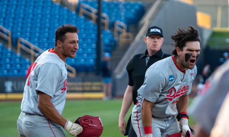 Two baseball players in gray uniforms celebrate on the field; one is holding a helmet and smiling, the other is shouting with black face paint. An NCAA official stands behind them. Blue stadium seats are visible in the background.