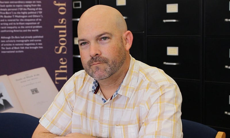 A man with a bald head and trimmed beard sits at a desk, wearing a plaid shirt. Filing cabinets and a poster with text are visible in the background. 