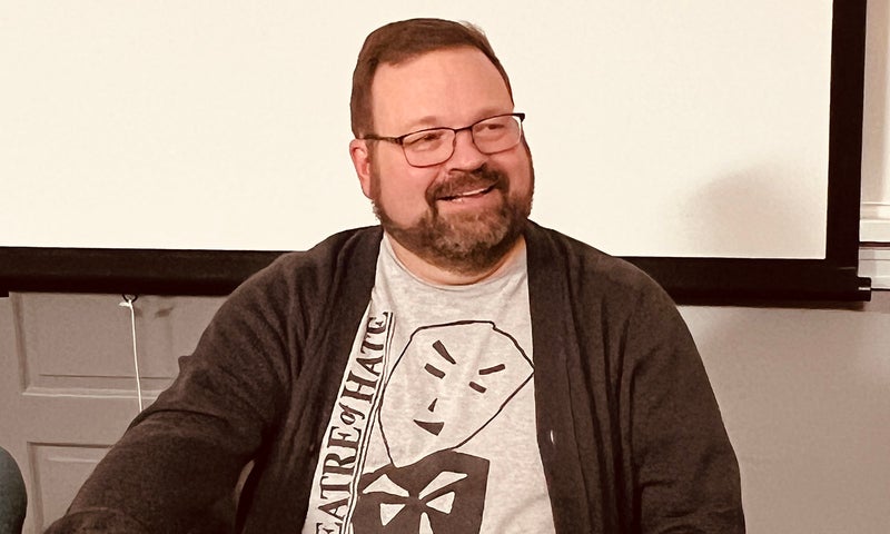 A man with glasses and a beard sits in a classroom engaged in conversation.
