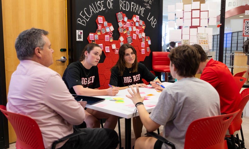 Four people sit around a small white table in discussion, with papers and markers on the table, in a classroom with red chairs and a wall.