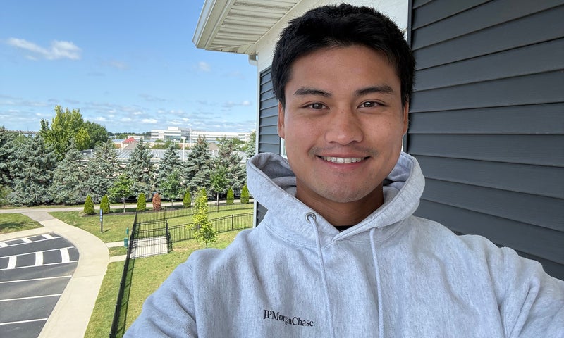 A person wearing a light gray hoodie smiles for a selfie outside near a building, with trees, grass, and a parking lot in the background under a blue sky.