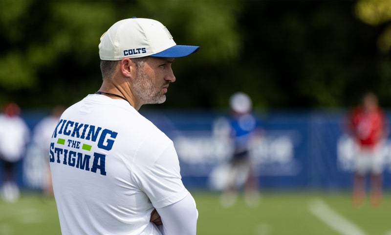 A man in a white Kicking the Stigma shirt and Colts cap stands on a sports field, watching an outdoor practice session.