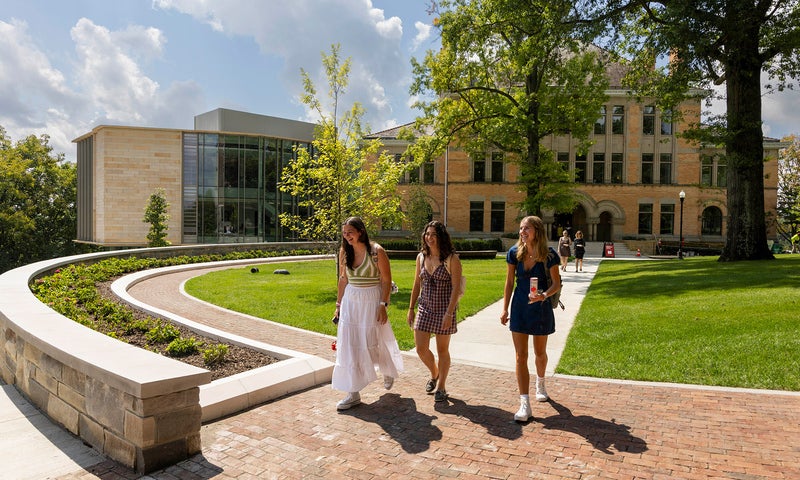 Three people walk along a brick path in front of a modern academic building on a sunny day, surrounded by green lawns and trees.