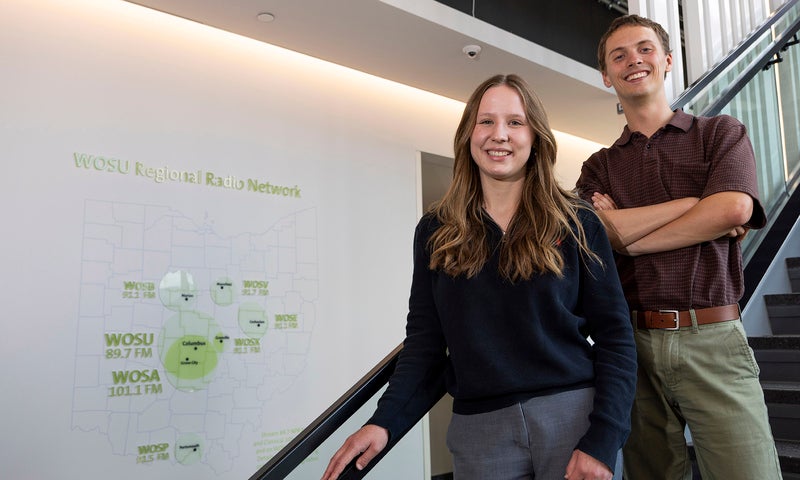 Two people stand side by side on a staircase indoors, smiling at the camera. A diagram is visible on the white wall behind them.