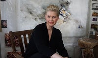 Allison Leigh, a woman with blonde hair, sits on a wooden chair in front of an abstract painting, wearing a black outfit. Artworks and materials are visible in the background.