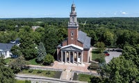A red brick church with a tall steeple, white columns, and a clock tower, surrounded by trees and a parking lot on a clear, sunny day.