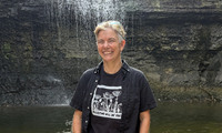 Person standing and smiling in front of a small waterfall, wearing a black shirt and sunglasses on their head.