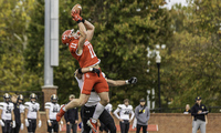 A football player in a red uniform jumps to catch a pass while a defender in a white and black uniform attempts to block him. Other players and trees are visible in the background.