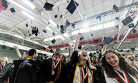 A large group of college graduates in caps and gowns toss their caps in the air in celebration in a large fieldhouse during a graduation ceremony.