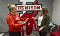 Two women stand indoors in front of a large Denison University logo. One hands a red sports jersey with the number 26 on it to the other. Both are smiling.