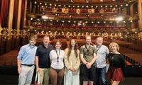 A group of seven people stand on a theater stage with rows of empty red seats and ornate balconies visible behind them.